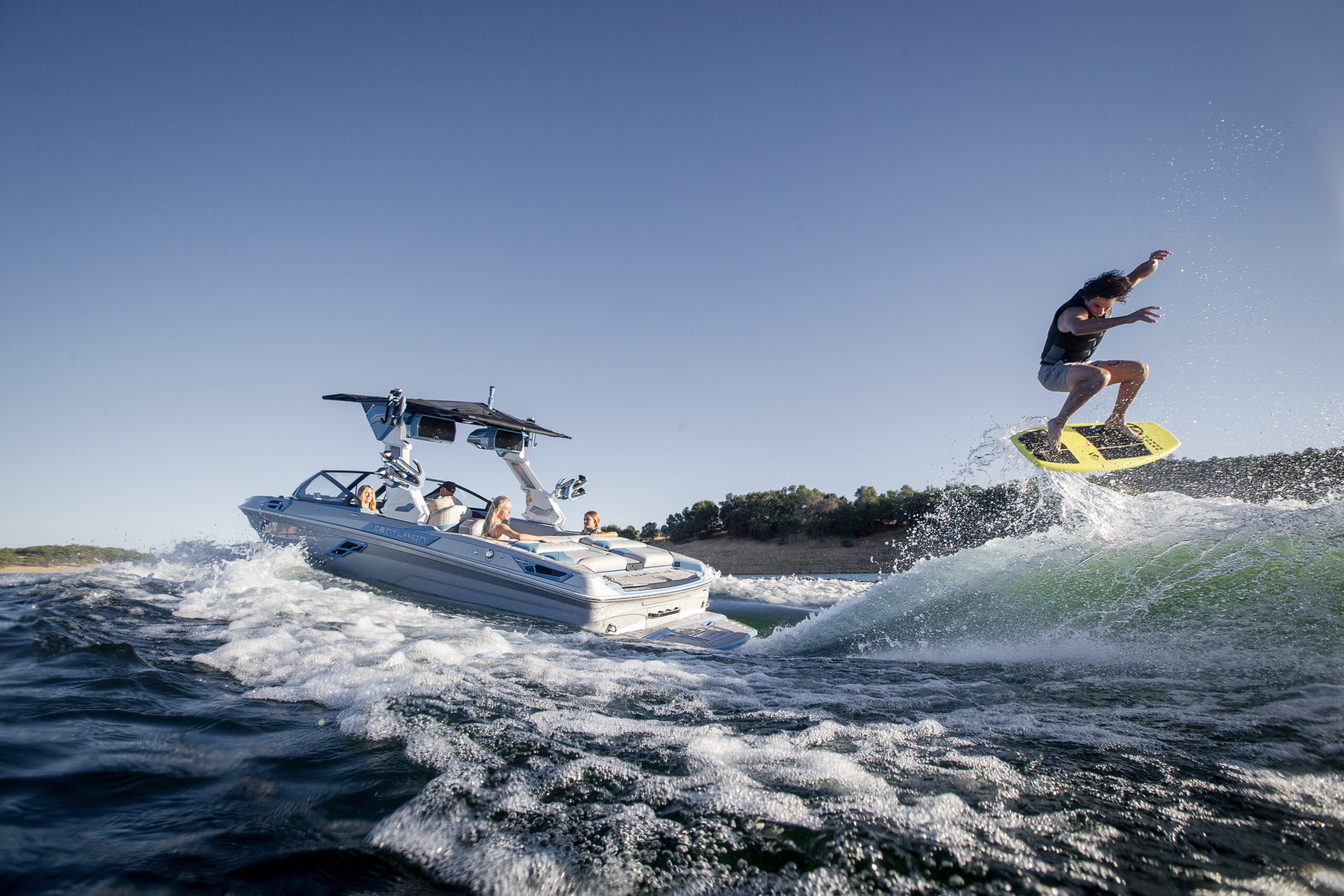 A person is wakeboarding on a wave behind a speedboat on a sunny day, with several people watching from the boat.
