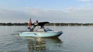 A light blue Centurion WSWS motorboat with an American flag moves across a calm lake under a partly cloudy sky, with the shoreline visible in the background at the Come and Take It Classic.