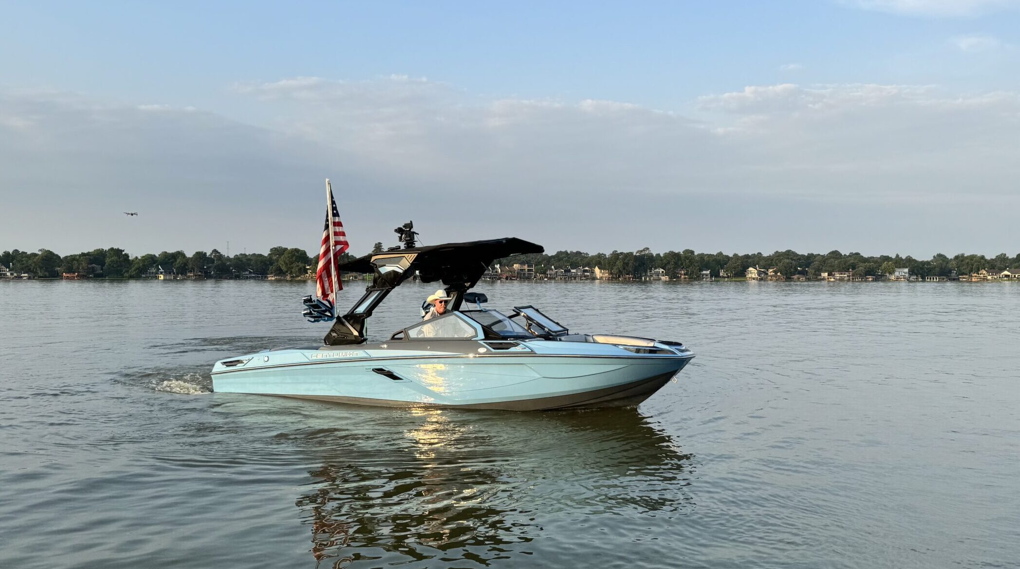 A light blue Centurion WSWS motorboat with an American flag moves across a calm lake under a partly cloudy sky, with the shoreline visible in the background at the Come and Take It Classic.