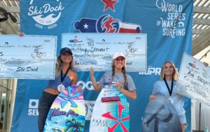 Three women holding large checks and surfboards stand atop the podium at the Centurion WSWS Come and Take It Classic wake surfing competition, with an event banner displayed in the background.