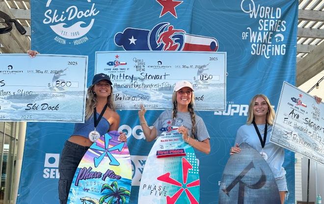 Three women holding large checks and surfboards stand atop the podium at the Centurion WSWS Come and Take It Classic wake surfing competition, with an event banner displayed in the background.