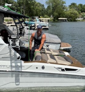 A man wearing a life jacket and cap stands on a docked boat, holding a rope, with docks, trees, and other boats visible in the background on a sunny Day 1 Recap of the SurfMN King of the Lakes Classic.