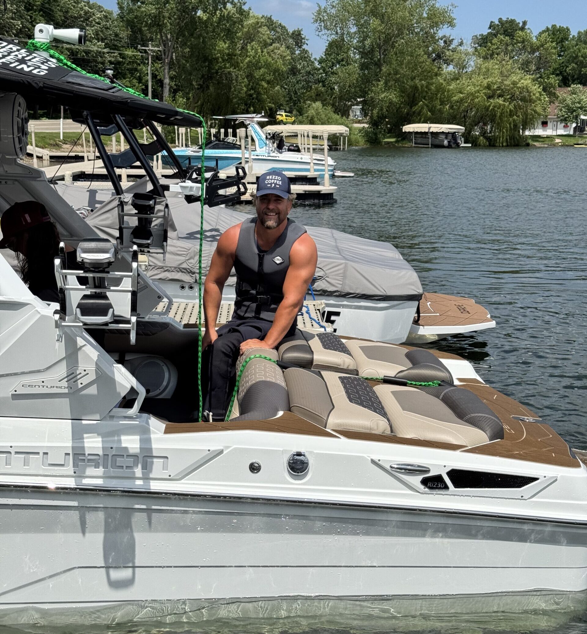 A man wearing a life jacket and cap stands on a docked boat, holding a rope, with docks, trees, and other boats visible in the background on a sunny Day 1 Recap of the SurfMN King of the Lakes Classic.