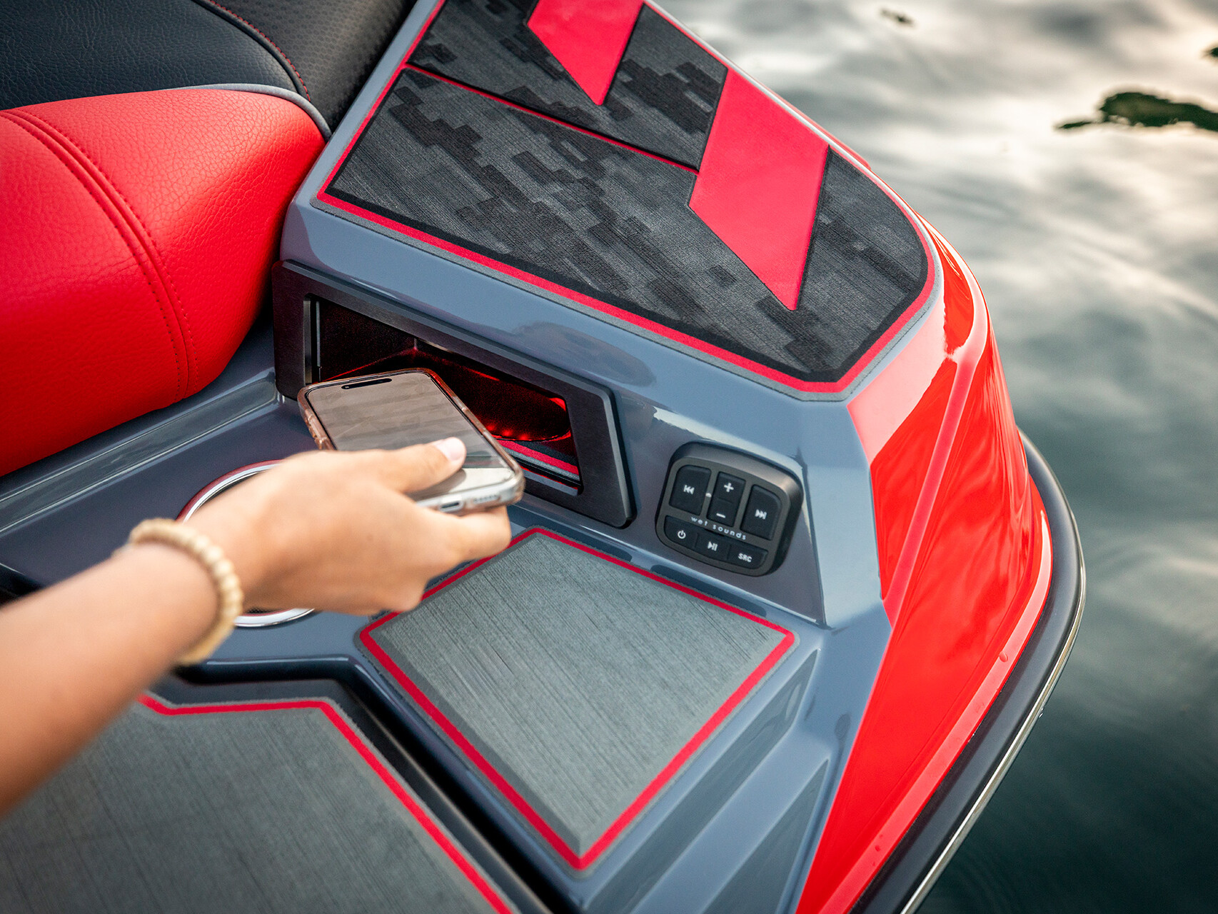 A person places a smartphone into a storage compartment on a red and gray Centurion Ri Series watercraft near the water’s edge.
