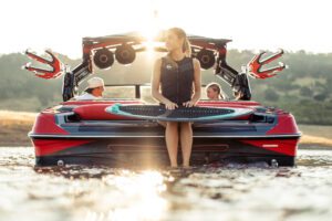 A woman sits on the back of a red Centurion Ri Series boat holding a wakeboard, while two people relax inside; the sun is low in the sky.