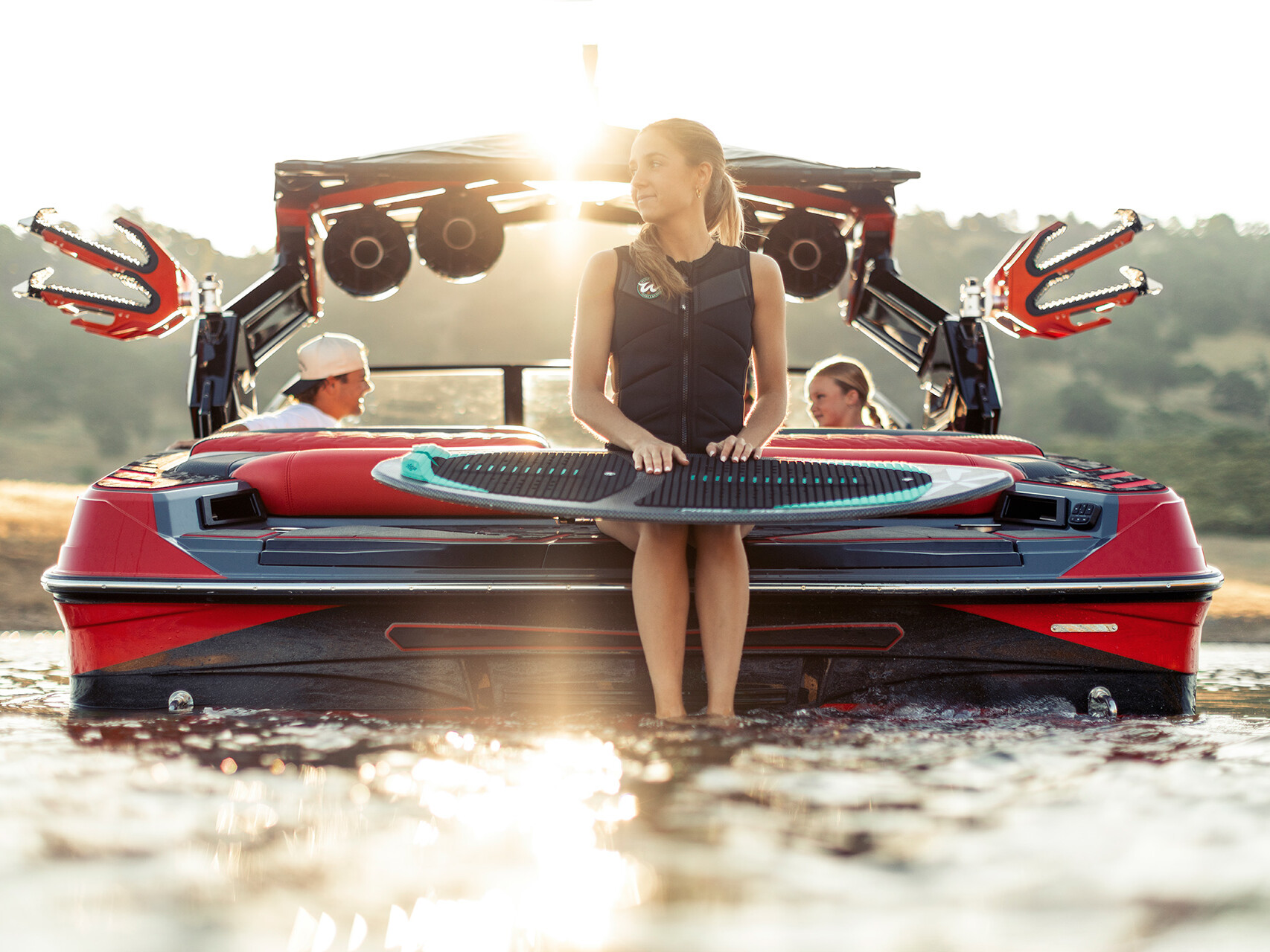 A woman sits on the back of a red Centurion Ri Series boat holding a wakeboard, while two people relax inside; the sun is low in the sky.