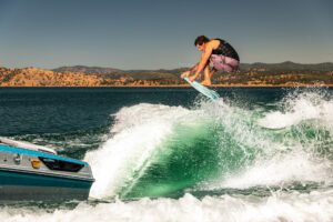 A person in a black vest and purple shorts performs a jump on a wakeboard behind a blue Centurion Ri Series boat on a sunny lake with hills in the background.