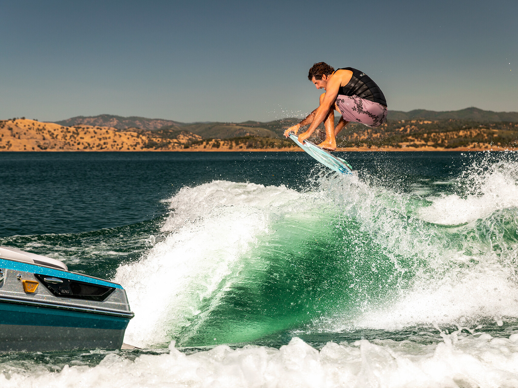A person in a black vest and purple shorts performs a jump on a wakeboard behind a blue Centurion Ri Series boat on a sunny lake with hills in the background.