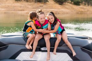 Three children wearing life jackets sit on the back of a Centurion Ri Series boat, smiling and hugging, with a lake and dry grassy shore in the background.