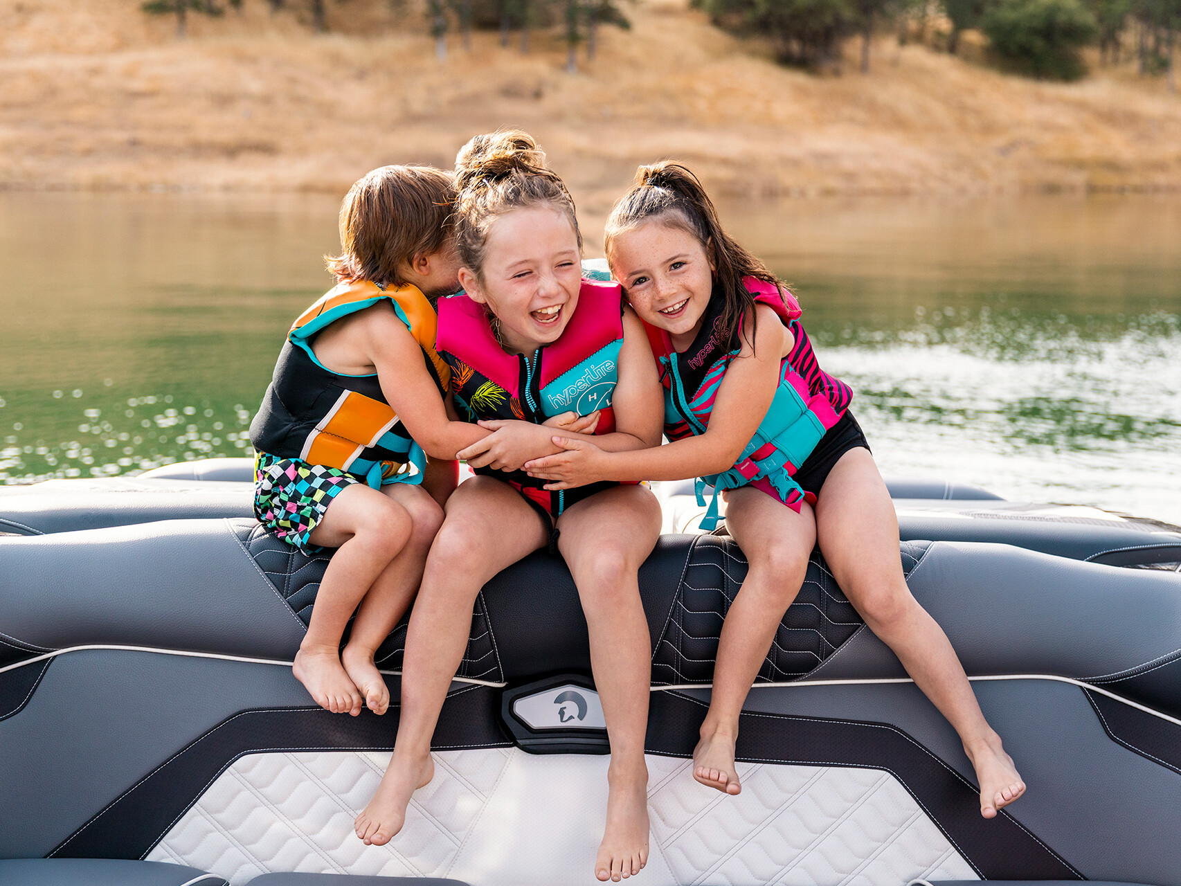 Three children wearing life jackets sit on the back of a Centurion Ri Series boat, smiling and hugging, with a lake and dry grassy shore in the background.