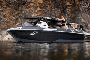 A sleek black and gray Centurion Ri Series boat with three people on board is seen on calm water near a rocky shoreline.
