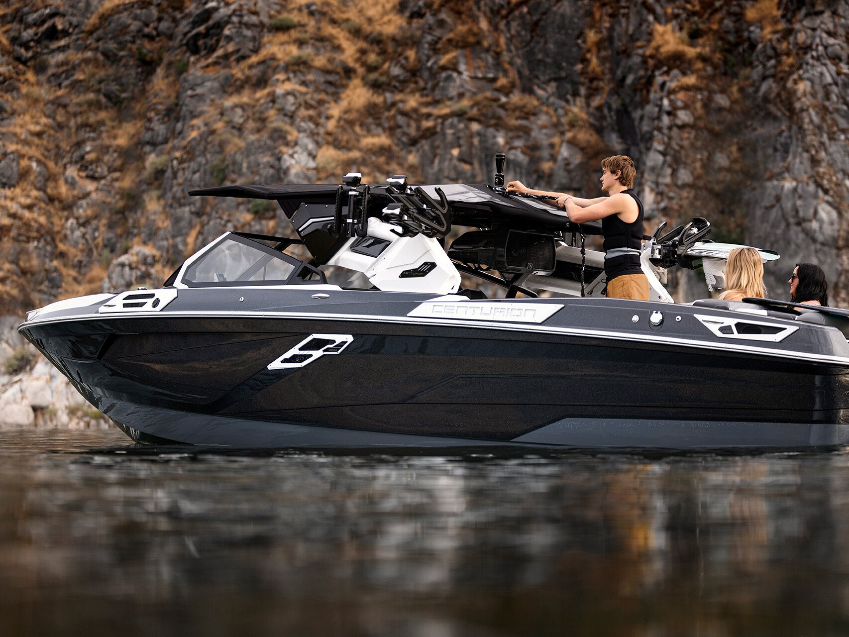 A sleek black and gray Centurion Ri Series boat with three people on board is seen on calm water near a rocky shoreline.