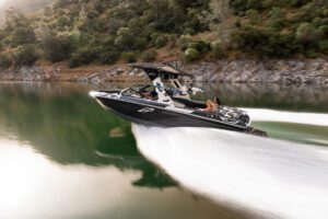 A Centurion Ri Series speedboat with several passengers moves quickly across a calm lake, creating a large wake, with forested hills in the background.