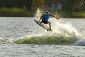 A person wearing a blue vest is jumping on a wakeboard above the water, creating a splash behind them on a lake.