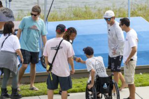 A group of people, including a man in a wheelchair, gather by a lakeside dock. Two men shake hands while others stand around them, talking and smiling.