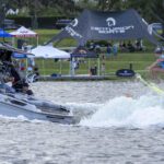 A person in a blue vest is wakesurfing behind a Centurion boat on a lake during the Wake Surfing Championship, with spectators and sponsor tents, including GM Marine, visible on the grassy shore.