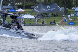 A person in a blue vest is wakesurfing behind a Centurion boat on a lake during the Wake Surfing Championship, with spectators and sponsor tents, including GM Marine, visible on the grassy shore.