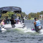 A group of people sit on a Centurion World boat, watching a man wakeboarding behind it, while another person is seen in the water nearby.