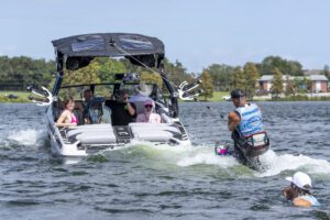 A group of people sit on a Centurion World boat, watching a man wakeboarding behind it, while another person is seen in the water nearby.