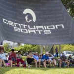 A group of people sits under a large canopy tent labeled "Centurion Boats" on a grassy area during the Wake Surfing Championship, with trees and cars visible in the background.