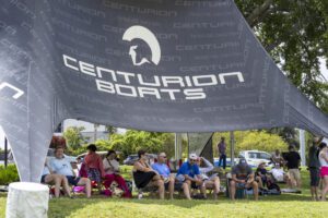 A group of people sits under a large canopy tent labeled "Centurion Boats" on a grassy area during the Wake Surfing Championship, with trees and cars visible in the background.