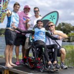 Five men pose together on a lakeside dock; one is in a wheelchair holding a paddleboard, while others stand beside him, smiling with surfboards—celebrating their time at the Centurion World Wake Surfing Championship.