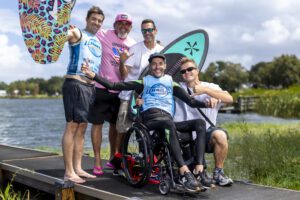 Five men pose together on a lakeside dock; one is in a wheelchair holding a paddleboard, while others stand beside him, smiling with surfboards—celebrating their time at the Centurion World Wake Surfing Championship.