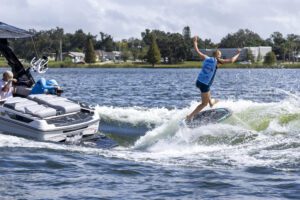 A person rides a wakeboard on a lake behind a boat, with arms raised, while people sit and watch from the boat.