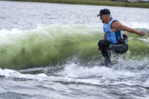 A man wearing a blue tank top and black cap wake surfs on adaptive equipment, riding a wave on a lake.