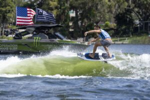 A person in athletic gear rides a wakeboard on a wave behind a boat displaying American flags and sponsor logos on a sunny day.