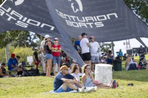 People sit and stand on a grassy area under a large "Centurion Boats" canopy at an outdoor event, with various chairs, bags, and coolers visible.