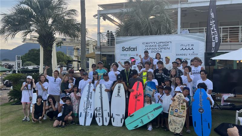 A group of people, some holding surfboards, pose for a group photo outdoors in front of a Centurion Boats banner at the 2024 Centurion Wake Surf Japan Open, part of the World Series of Wake Surfing Japan Tour.