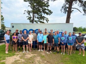 A group of people stand together outdoors in front of a white trailer with "Operation" and "Outfit" visible on its side at an S2S Event near Lake Conroe. Trees and grass provide a scenic backdrop.