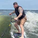 A man with tattoos and a life vest rides an electric surfboard on Lake Conroe, holding a handle attached to a rope, with waves and a blue sky in the background during the S2S Event.