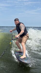 A man with tattoos and a life vest rides an electric surfboard on Lake Conroe, holding a handle attached to a rope, with waves and a blue sky in the background during the S2S Event.