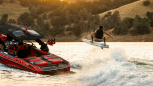 A person on a wakesurf board catches air behind a red tow boat on a lake, with hills and trees in the background at sunset.
