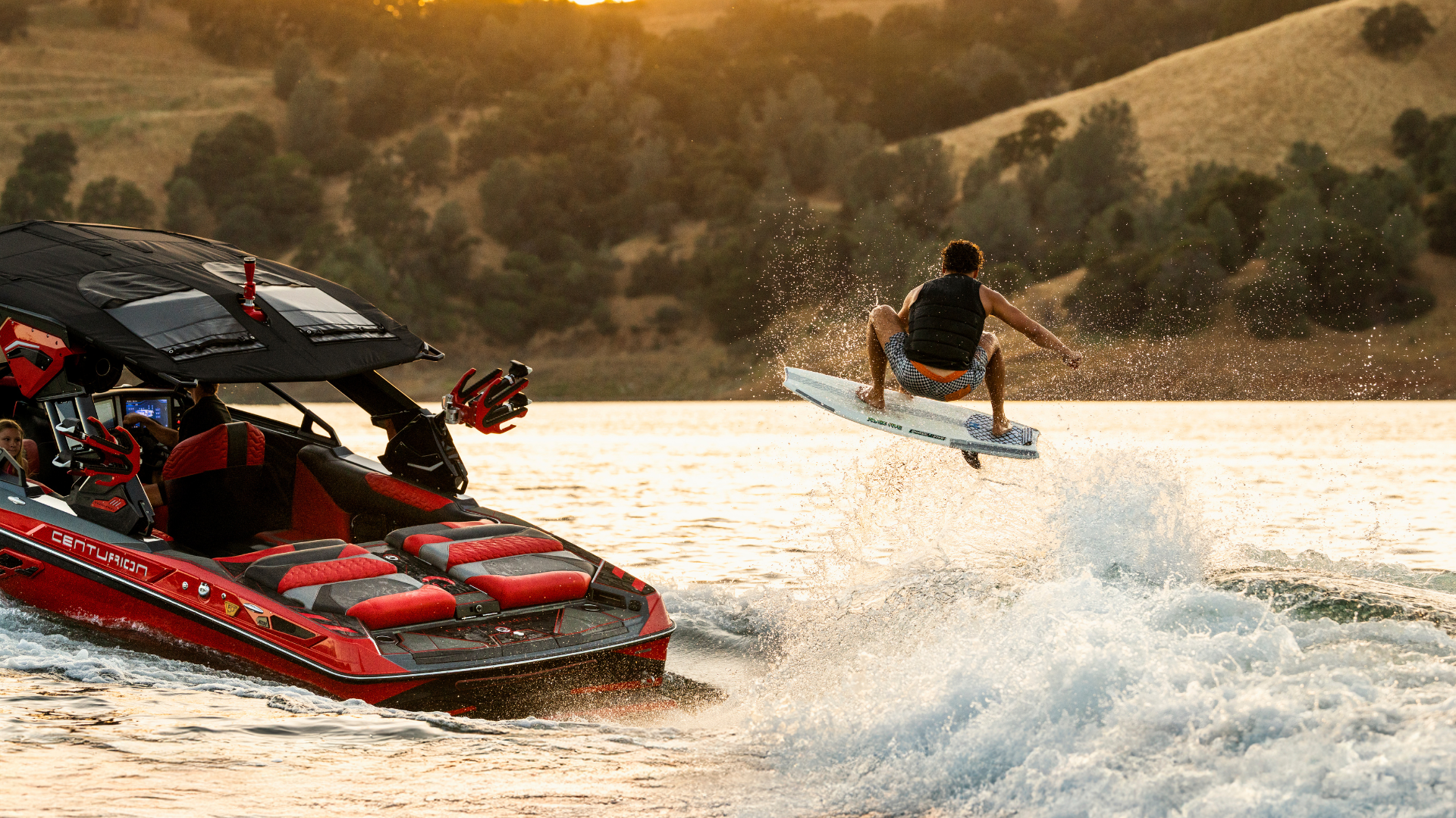 A person on a wakesurf board catches air behind a red tow boat on a lake, with hills and trees in the background at sunset.