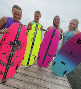Four young girls stand on a dock, each holding a colorful wakesurf board and smiling at the camera under a cloudy sky, ready for the Conroe Classic with Centurion Boats in the background.