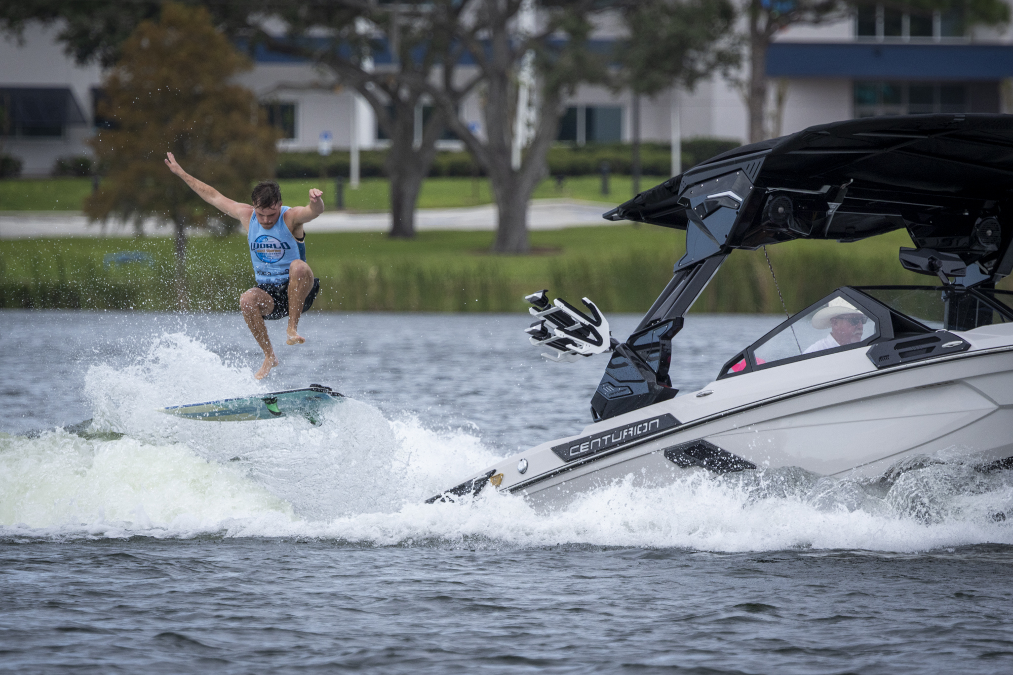 A person in a blue tank top jumps off a wakeboard behind a white Centurion boat at the Centurion World Series on a lake, with trees and buildings in the background.
