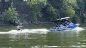 A person wakeboarding on a lake is being towed by a blue and white Centurion Boat from CBK Watersports, with three people riding in the boat and trees visible on the shore in the background.