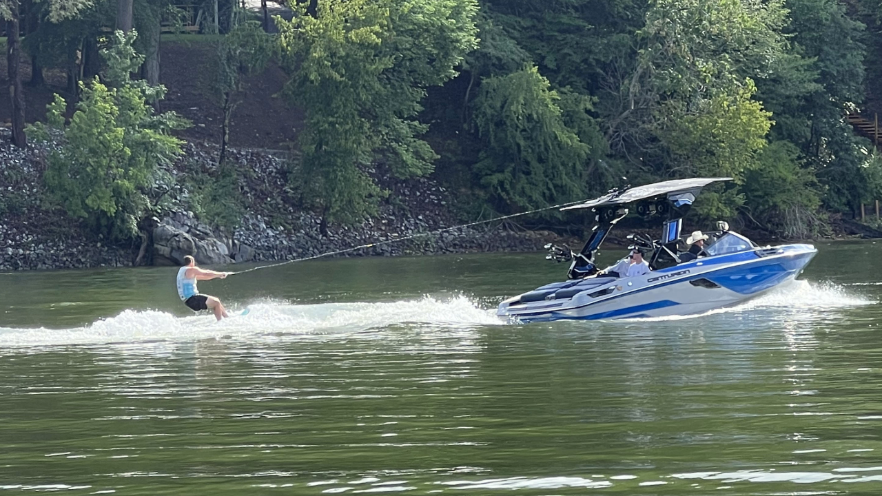 A person wakeboarding on a lake is being towed by a blue and white Centurion Boat from CBK Watersports, with three people riding in the boat and trees visible on the shore in the background.
