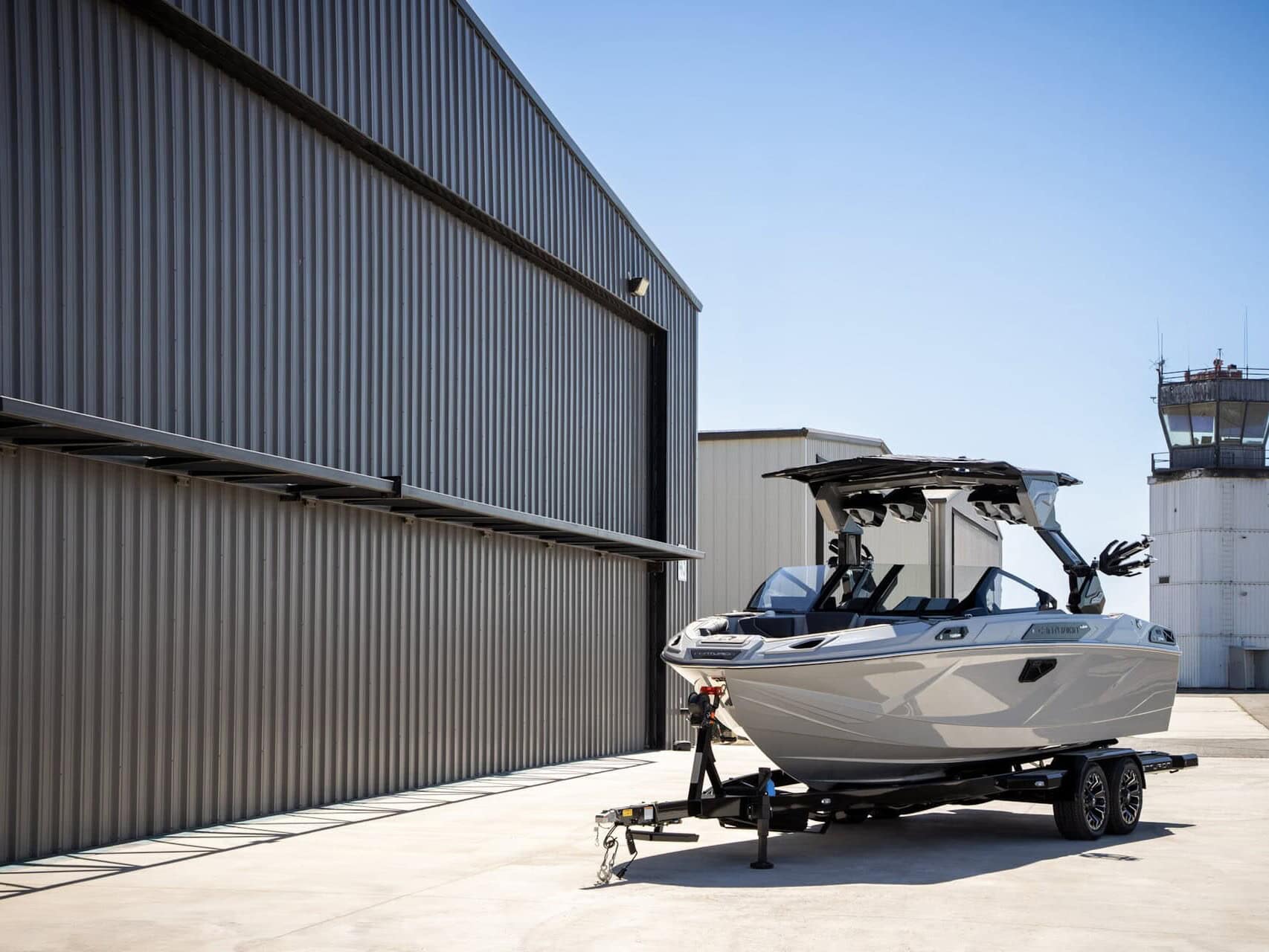 A white Centurion Ri Series boat on a trailer is parked beside a large metal building near an air traffic control tower under a clear blue sky.