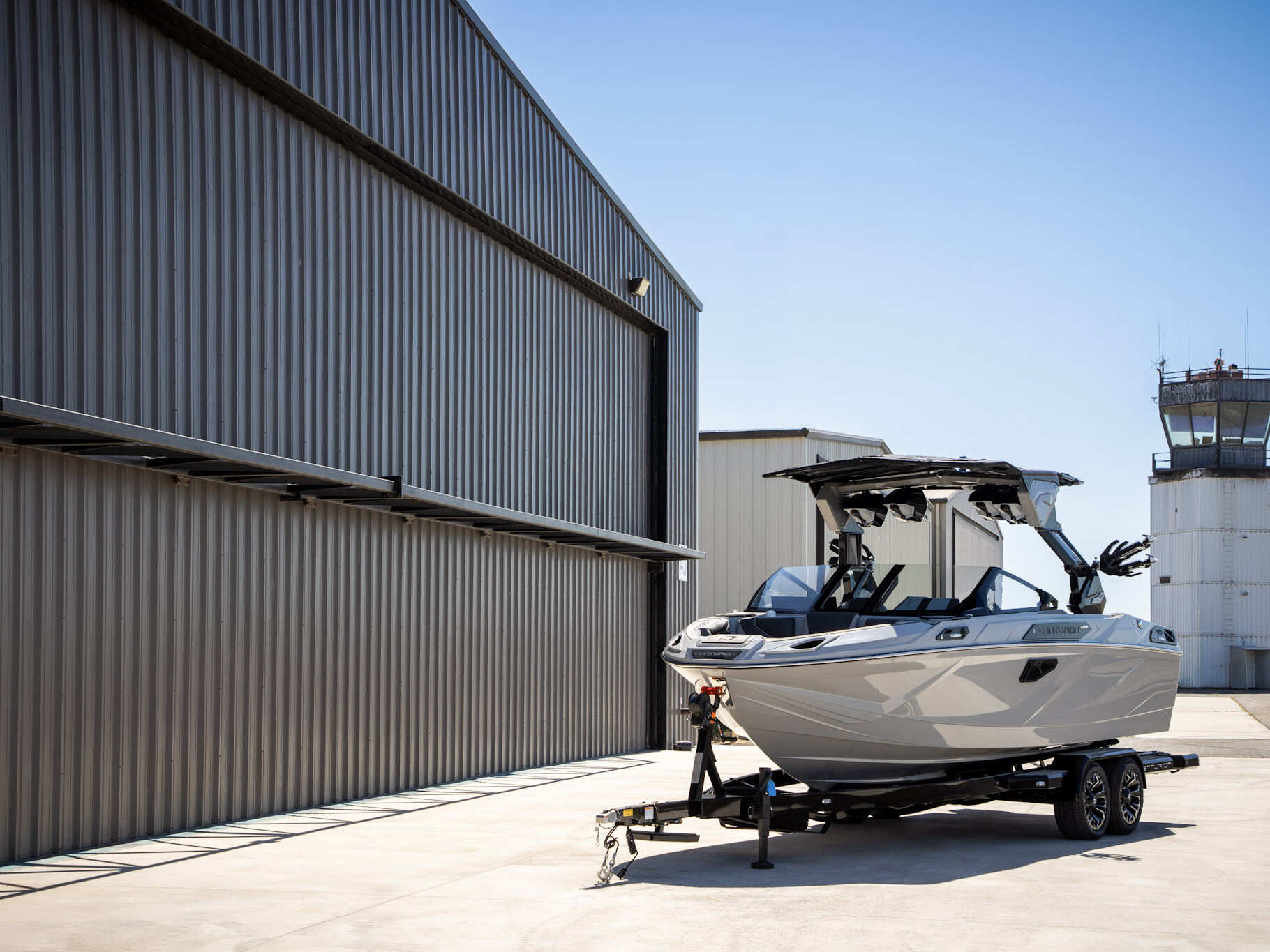 A white Centurion Ri Series boat on a trailer is parked beside a large metal building near an air traffic control tower under a clear blue sky.