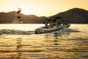 A person wakesurfs behind a Centurion Ri Series boat at sunset, performing a backflip over the water with mountains visible in the background.