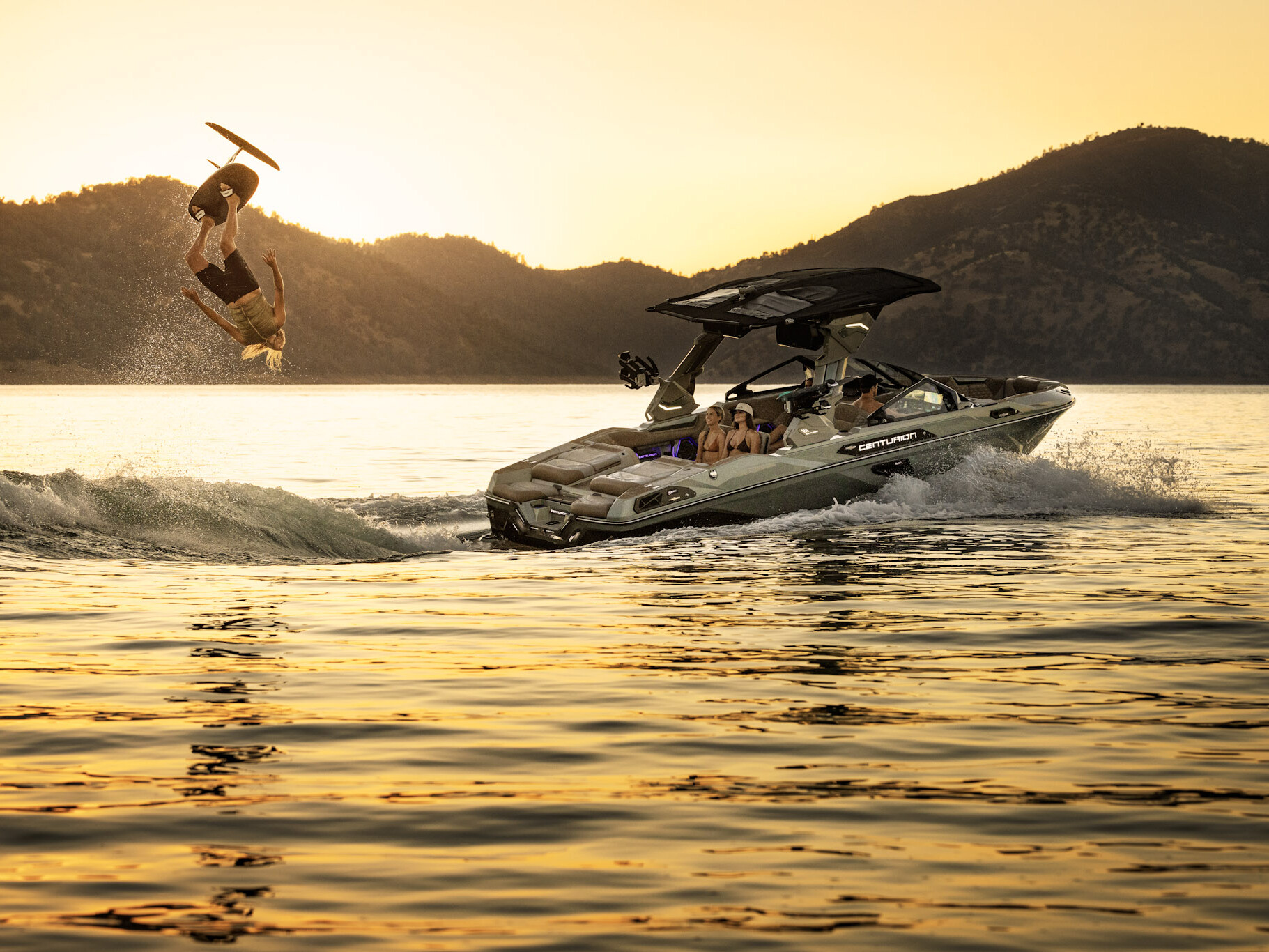 A person wakesurfs behind a Centurion Ri Series boat at sunset, performing a backflip over the water with mountains visible in the background.