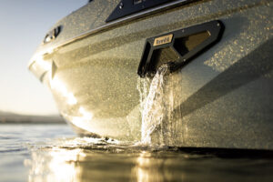 Close-up of water pouring out of a Centurion Ri Series boat’s ballast vent labeled "Rawfill," with sunlight reflecting off the boat’s hull at the water’s surface.