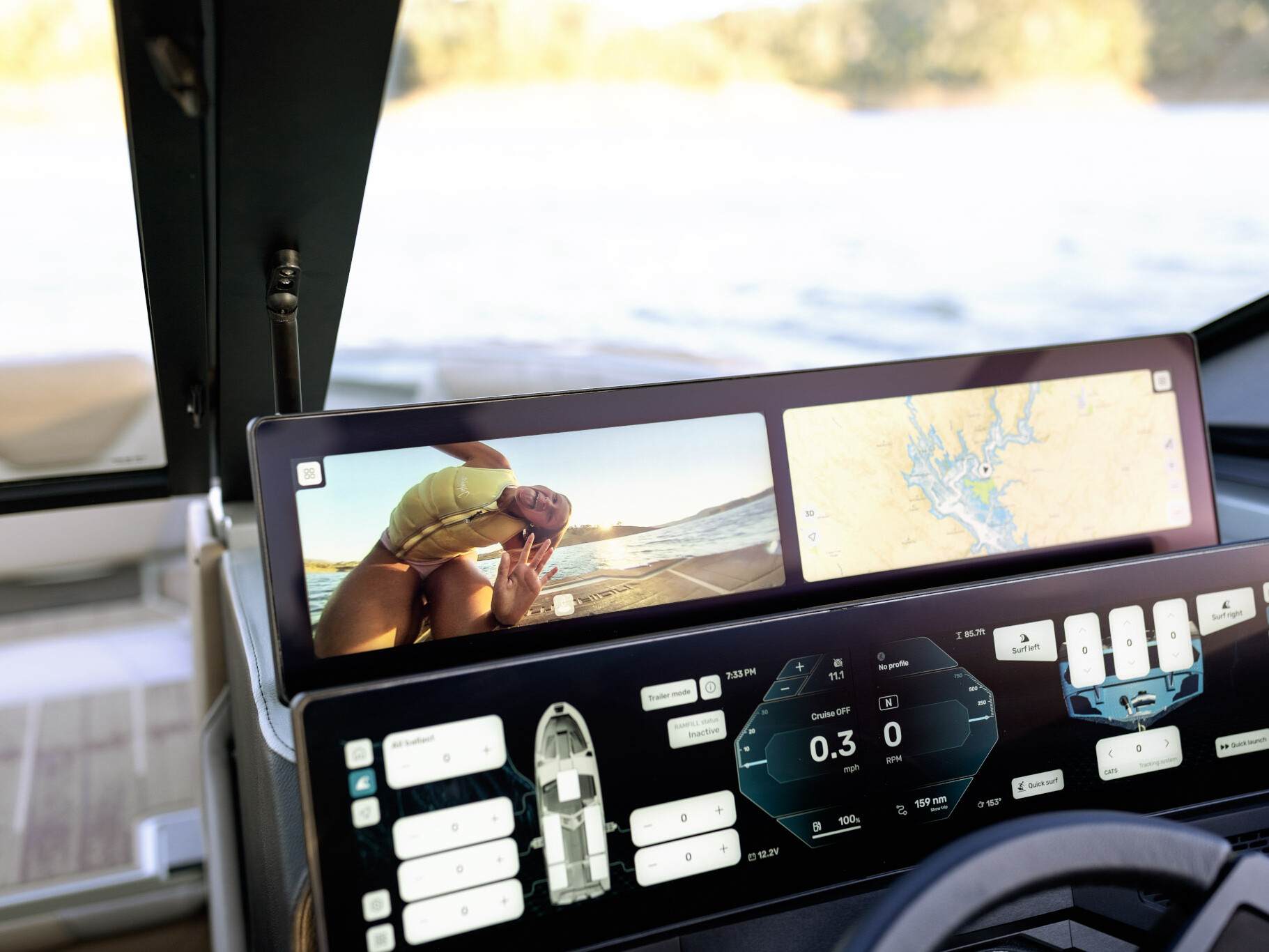 Dual digital screens on The Centurion Ri Series boat's dashboard display a map, boat controls, and a video of a person in swimwear on a beach. Water and part of the boat deck are visible in the background.