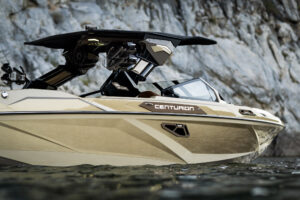 A beige Centurion Ri Series speedboat floats on calm water near a rocky shoreline, with the boat’s black roof and interior details visible.