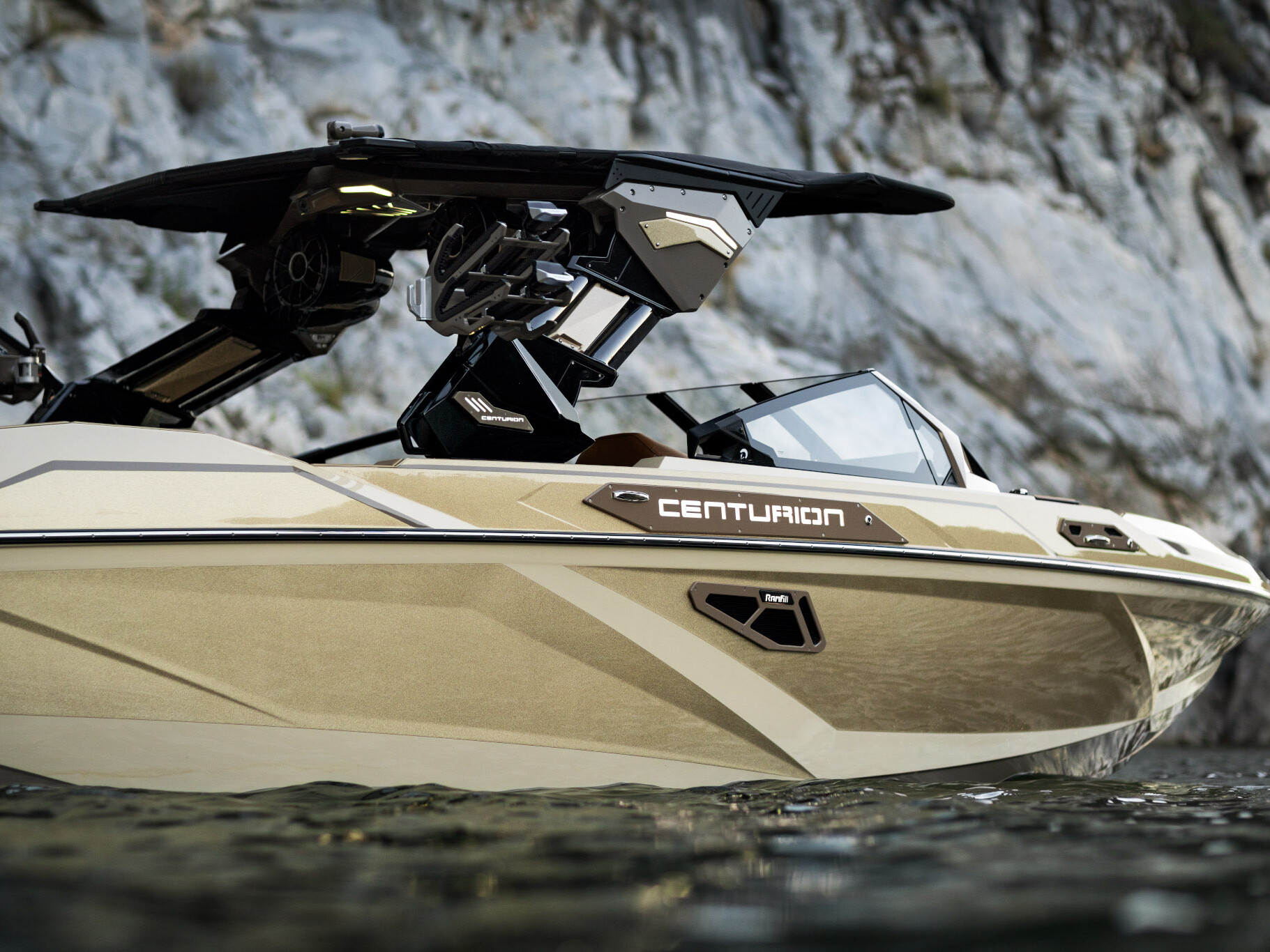 A beige Centurion Ri Series speedboat floats on calm water near a rocky shoreline, with the boat’s black roof and interior details visible.
