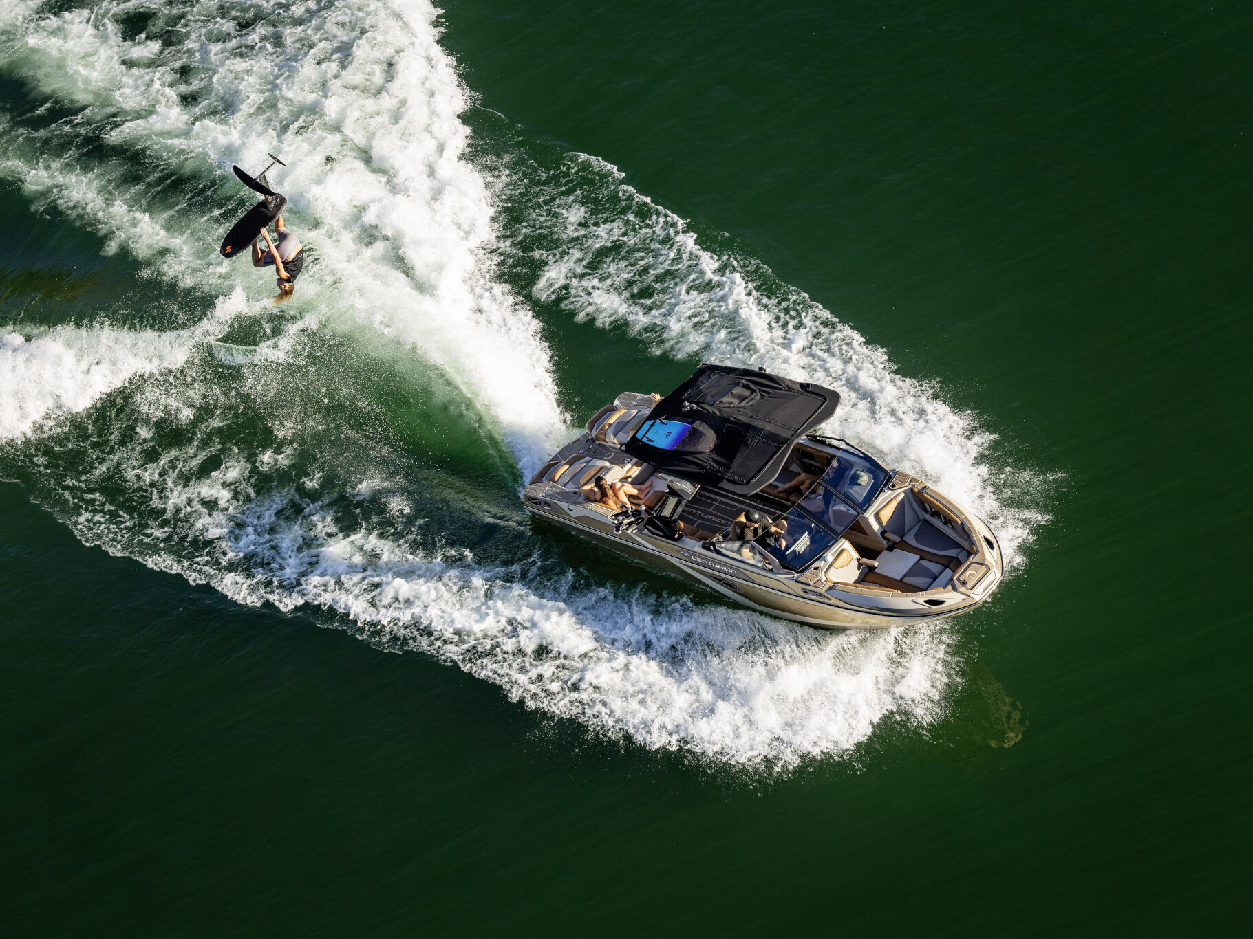 A person wakeboards behind a motorboat, possibly from The Centurion Ri Series, on green water, leaving curved white waves in their wake.